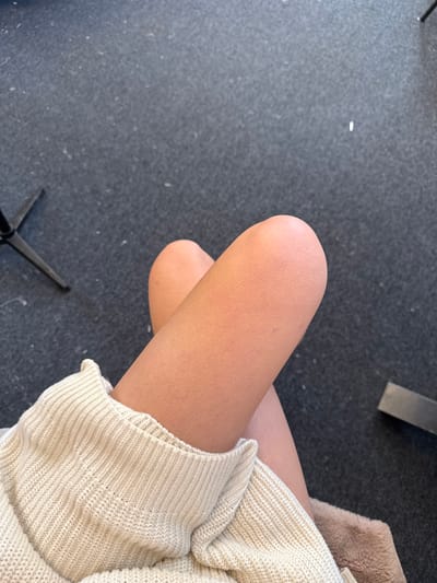 The image displays a first-person perspective view of an individual's crossed legs within an indoor setting in Arcueil, France. The legs, exhibiting a light to medium skin tone with visible hair follicles, are partially covered by a light beige or cream-colored ribbed knit sweater on the lower left. A section of light brown, textured material, possibly faux-fur or plush fabric, is visible beneath the legs on the lower right. The background consists of a dark grey carpeted floor with scattered light-colored specks. In the upper left quadrant, a portion of a black, three-legged object, consistent with a tripod or stand base, is present. A dark, rectangular object lies on the carpet in the lower right background.