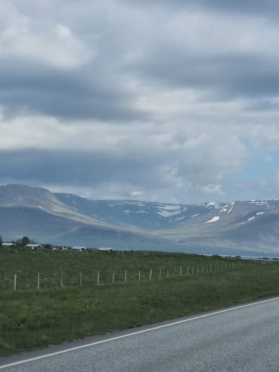 The image depicts a scenic view of the Icelandic landscape, likely near Akranes. In the foreground, a paved road with a white line marking the edge runs diagonally from the bottom right corner towards the left. Adjacent to the road is a grassy field with a simple fence composed of wooden posts. In the middle ground, a row of small, white-roofed buildings is visible on a slightly elevated grassy area. Beyond these buildings, rolling hills rise, culminating in a majestic mountain range. The mountains are partially covered in snow, with stark white patches contrasting against the muted greens and grays of the slopes. The sky above is overcast with heavy, grey clouds, suggesting a cool, possibly misty, atmosphere. The overall impression is one of a rural, somewhat isolated, yet beautiful natural environment.