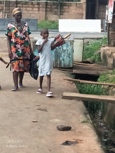The image depicts an outdoor scene in Akpuru, Nigeria. On the left, an adult woman with dark skin and a patterned headwrap wears a colorful floral short-sleeved dress in shades of green, orange, pink, black, and white, with a V-neck. She wears light blue sandals and carries a black bag in her left hand. To her right, a child with short hair wears a light grey short-sleeved top over a pleated light blue underskirt, paired with pink and white striped slide sandals. This child holds a folded, dark red-brown umbrella with a wooden handle in their left hand and has their right hand positioned at their mouth. A partial view of a third, smaller figure wearing a yellow top is visible on the far left.

The ground consists of an uneven, light brown dirt path. A small, flat, metallic, coin-like object and a dark rock are visible on the path. A dark, irregular stain is also present on the ground. Along the right side of the path runs an open drainage ditch containing stagnant water, various pieces of litter, and green vegetation. Two rough wooden planks are laid across this ditch, forming makeshift bridges.

In the middle ground, situated behind the walking figures and near the ditch, are two containers: a rusted blue metal barrel and a lighter-colored, possibly grey, rectangular metal container. Behind these objects, a paved road is visible, bordered by a white concrete element. Further in the background, a white rectangular concrete barrier stands in front of a building constructed from light brown concrete blocks, featuring a dark, open doorway. Beyond this, additional block walls and green foliage are present. A partially visible poster with red text on a white background is affixed to a pole on the far right of the image.