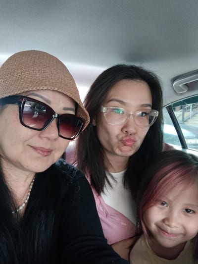 The image shows a selfie taken inside a vehicle, likely on a street in Yahşihan, Turkey, given the context. Three people are visible, appearing to be a family. In the foreground, on the left, a woman wearing a straw hat and large sunglasses smiles at the camera. She has on a black top and a pearl necklace. To her right and slightly behind her is another woman wearing glasses and making a duckface. She has dark hair and is wearing a pink shirt. In front of her and to the right is a young girl with pink-streaked hair, who is smiling at the camera. The interior of the car, including the headliner and a portion of a window, is visible in the background. The lighting suggests it is daytime.