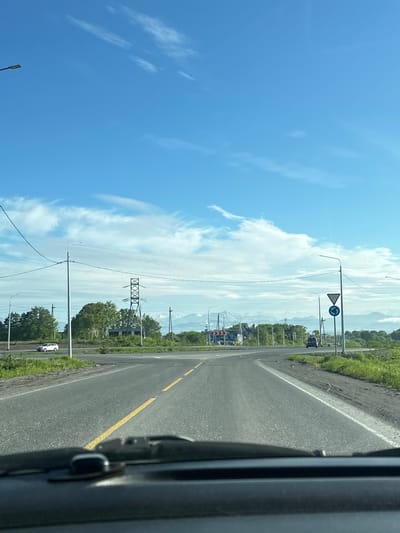 A daytime, eye-level, wide shot captures a view from the perspective of a vehicle's dashboard looking out at a rural road intersection under a bright blue sky with scattered white clouds. The road is paved and has a yellow dividing line. Several cars, a white SUV and a dark-colored SUV, are visible on the road. There are utility poles and power lines crossing the frame. In the distance, a tall lattice transmission tower and a cluster of trees are present. Beyond the trees, snow-capped mountains are visible under a hazy sky. Road signs, including a yield sign and a roundabout sign, are posted at the intersection. The foreground shows the vehicle's dark-colored dashboard and windshield wiper. The overall impression is that of a scenic, rural landscape during daylight. The image is not specific to the city of Svetly, Russia; it appears to be a more general rural scene.