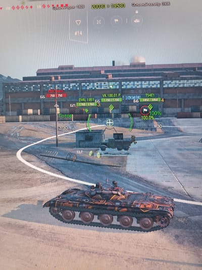 The image depicts a scene from a video game, likely World of Tanks, showing a battle scenario. The foreground features a camouflaged tank with orange and black tiger stripes on a dirt and asphalt surface. In the midground, there is a building complex with windows and industrial elements, as well as other tanks and game interface elements such as health bars, player names, and aiming reticles. The background shows a sky and distant landscape. The user has requested that this scene be described as if it were the city of Kurchaloy, Russia, which is not represented in the image.