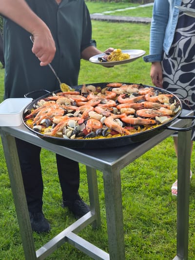 The image shows a close-up of a large paella dish filled with seafood, including shrimp, mussels, and clams, all mixed with yellow rice. The paella is situated on a metal table outdoors, with a grassy lawn visible in the background. A person on the left, wearing a dark green shirt and black pants, is using a silver serving spoon to scoop food from the paella. To the right, a person wearing a blue denim jacket and a black and white patterned dress is visible, also holding a plate with some paella on it. The setting appears to be a garden or outdoor event space. The presence of paella, a Spanish dish, and the outdoor setting suggest a social gathering or catering event. The image does not contain specific landmarks to pinpoint the location within Bettembourg, Luxembourg.