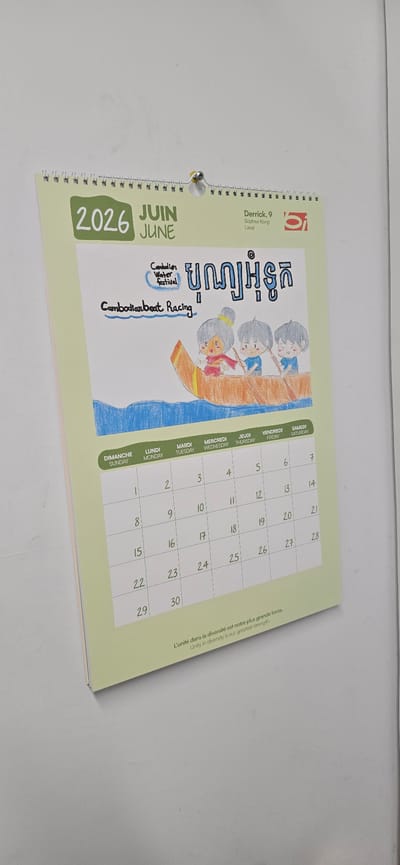 This is a close-up, slightly angled shot of a calendar page for June 2026. The page features a whimsical, hand-drawn illustration of three children in a boat, likely participating in a "Cambodian Water Festival" or "Cambodian Boat Racing" event, as indicated by the text. The drawing style is simple and colorful, with the children depicted with large eyes and cheerful expressions. The text "បុណ្យអុំទូក" in Khmer script is prominent at the top of the illustration.
To the left of the illustration, the calendar month is clearly marked as "JUIN JUNE" and the year "2026." A small detail on the upper left indicates "Derrick, 9 Sophea Kong Laval," possibly a dedication or the name of the artist.
The right side of the page shows a standard grid calendar layout for June. The days of the week are listed in French and English: DIMANCHE SUNDAY, LUNDI MONDAY, MARDI TUESDAY, MERCREDI WEDNESDAY, JEUDI THURSDAY, VENDREDI FRIDAY, SAMEDI SATURDAY. The dates are numbered sequentially from 1 to 30.
At the bottom right, a motivational quote appears in both French and English: "L'unité dans la diversité est notre plus grande force. Unity in diversity is our greatest strength." The overall impression is bright and positive, celebrating a cultural event through children's art. The setting appears to be indoors, likely a wall where the calendar is hung.