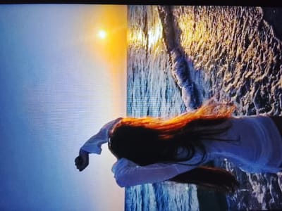 The image displays a woman with long, reddish-brown hair lying on what appears to be a rocky shore with waves crashing. She is wearing a white, long-sleeved shirt. The sun is setting or rising, casting a warm orange glow on the water and rocks. The background is a gradient of blue and orange, suggesting a sky and ocean. There is no indication of the city of Safonovo, Russia, or any urban elements present in the scene.