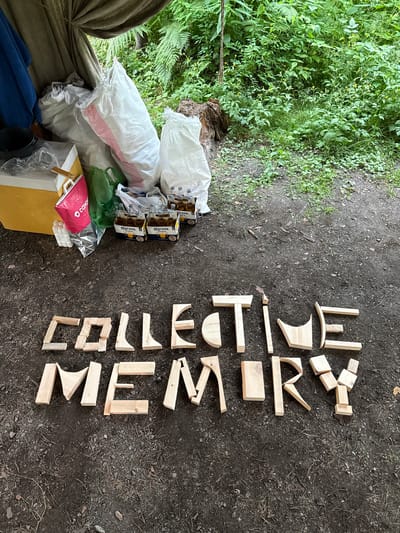 In Cheposh, Russia, outdoors on a dirt ground, wooden blocks are arranged to spell out "COLLECTIVE MEMORY". In the background, a canopy made of olive-green fabric is partially visible, with lush green foliage and ferns behind it. To the left, several large white sacks are piled up, alongside a yellow cooler, a pink shopping bag, and several cases of Corona beer.