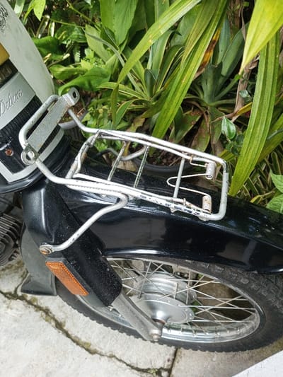 The image depicts a close-up, outdoor view of the front section of a motorcycle, stationary on a concrete surface in Salai Har Htan, Myanmar. The motorcycle's front is primarily black, with a lighter upper panel displaying "Deluxe" and partially visible "ZUKI" branding. A silver metal front rack, exhibiting wear and minor corrosion, is mounted above the black front fender and features two coiled springs on its right side. An orange rectangular reflector is affixed to the lower left fender. The front wheel, partially visible at the bottom, features a silver rim, spokes, and a tire with a distinct tread pattern. Metallic components of the front suspension and engine casing are visible on the left. The background is composed of dense, lush green foliage with various plant types, some showing signs of discoloration, and a partially visible black plant pot.