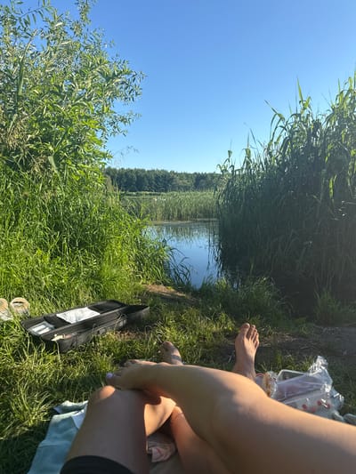 The image depicts a ground-level view of a peaceful outdoor scene, likely in the vicinity of Zaścianki, Poland, given the presence of lush vegetation bordering a body of water. Two individuals are partially visible, their legs and feet positioned in the foreground, suggesting they are relaxing on the grassy bank. One individual's bare feet are prominent, while the other's legs are also in view, resting near a light blue towel.
In the middle ground, a black case containing what appears to be fishing equipment or tools is placed on the grass, alongside a clear plastic bag containing food items, possibly a sandwich and berries.
The environment is characterized by abundant green grass and tall reeds or rushes along the edge of a calm body of water, which reflects the clear blue sky. A dense line of trees forms the horizon in the distance, with more reeds visible on the far side of the water. The overall atmosphere is serene and suggestive of a recreational outing by the water.