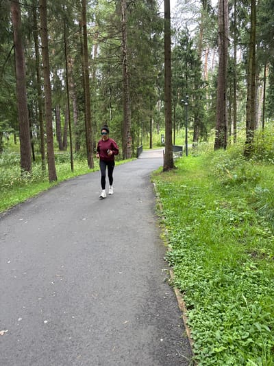A woman is captured mid-stride, jogging on an asphalt path through a dense forest. She is wearing a maroon long-sleeved shirt, black leggings, and white athletic shoes. She has sunglasses on and her hair is pulled back. The path is bordered by lush green grass and numerous tall, slender pine trees. In the background, a streetlamp and a park bench are visible. The scene is likely located in Pavlovskaya Sloboda, Russia, given the characteristic flora of the region.