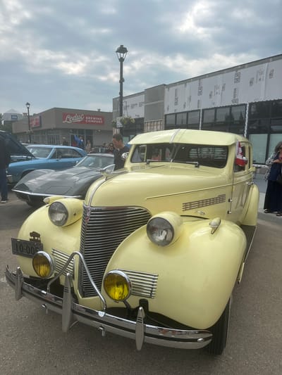 This media file captures a portion of a car show, likely in Leduc, Alberta, Canada, based on the visible signage. The main subject is a light yellow vintage car, prominently displayed at the forefront. Its chrome details, including the grille, headlights, and bumper, gleam. The car is adorned with a small Canadian flag on its driver's side.
Behind the vintage car, other vehicles are visible, including a dark gray car and a blue car, suggesting a line-up typical of car shows. In the background, a building with "Leduc Brewing Company" signage is clearly visible, indicating the location. The building appears to be under some construction or renovation, with materials like "TITAN" and "HAYDUK HARDWARE" visible on sheeting covering parts of the facade.
Several people are scattered around the scene, some looking at the cars, others conversing. The overall atmosphere appears to be casual and leisurely, fitting for an outdoor event. The sky is cloudy, suggesting overcast weather, and the lighting indicates it is likely daytime. The presence of streetlights and the type of buildings imply an urban or town setting. A license plate on the yellow car reads "ALBERTA 10-005 EXPIRES MARCH," providing specific location and temporal context.