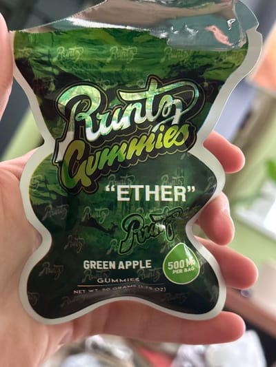 The scene depicts a close-up view of a human hand holding a green, irregularly shaped, soft-sided plastic pouch. The pouch, designed in the outline of a stylized character, features a white border and a dark green background with a holographic or reflective texture, patterned with repeated "Runtz" logos. Prominently displayed text on the pouch includes "Runtz Gummies" in large, stylized, iridescent lettering, followed by ""ETHER"" in white capital letters. Below this, "GREEN APPLE GUMMIES" is printed, with "NET WT. 50 GRAMS (1.76 OZ)" beneath. A green, leaf-shaped icon in the lower right corner displays "500 MG PER BAG". A slight tear or crease is visible on the top-left edge of the pouch. The hand, with visible skin tone and partial fingers, grasps the pouch. The background is blurred, indicating an out-of-focus indoor environment. The scene is located in the city of Dorchester, United Kingdom.