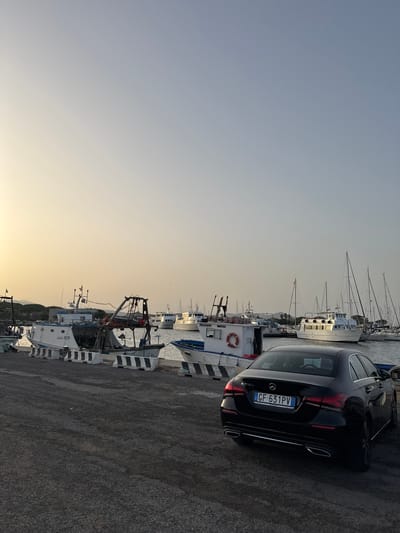 A black Mercedes-Benz A200d sedan is parked on a gravel surface in a harbor. The car's license plate reads "GF 631PV". In the background, several boats are docked, including fishing vessels and sailboats. Some of the fishing boats have visible names and numbers, such as "Adele 303229" and "PELICANO 304123". The water in the harbor appears calm. The sky is clear with a gradient of light blue and a soft yellow hue near the horizon, suggesting either sunrise or sunset. The scene is set in the vicinity of Thiniscole/Siniscola, Italy, a coastal town known for its fishing industry.