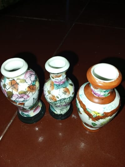 This image depicts three small, ornamental porcelain vases positioned on a dark brown, possibly tiled, surface in Longmiang, Laos. From left to right: The leftmost vase features a globular body with a flared rim, adorned with white and multi-colored floral patterns including pink, orange, and gold accents, resting on a black circular base. The middle vase is slender with a similar flared rim, decorated with white and multi-colored floral designs in pink, orange, and green, also on a black circular base. The rightmost vase has a broader body, a narrower neck, and an orange-brown rim, featuring white with multi-colored floral patterns including pink, yellow, and green, interspersed with orange-brown horizontal bands. All vases exhibit a glazed finish. The background is dark and indistinct. No individuals are present.