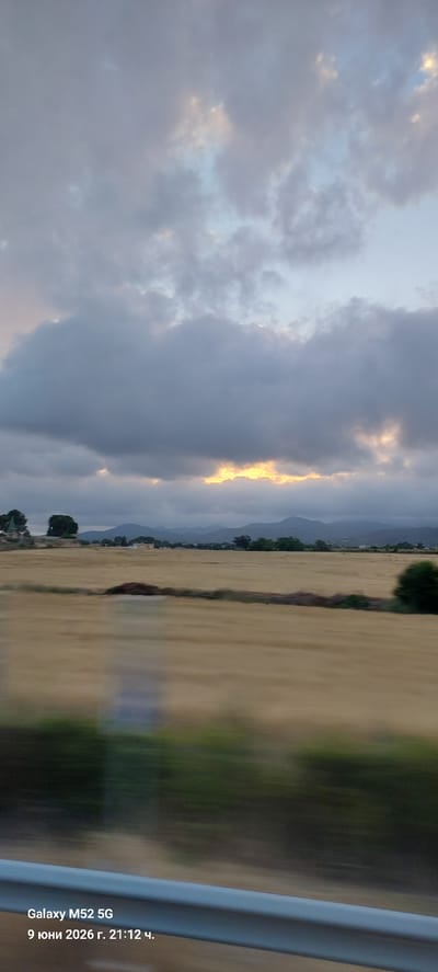 A blurred, motion-affected photograph captures a rural landscape under a cloudy sky. The sky is dominated by heavy, grey clouds with patches of illuminated sky showing through, suggesting either sunrise or sunset. The middle ground features a wide, golden field, possibly harvested wheat or dry grass, with distant, hazy mountains forming the horizon. A line of trees and some structures are visible along the edge of the field. The foreground shows a blurred streak of green, indicating vegetation, and a grey metallic rail, likely from a vehicle. In the bottom left corner, overlaid text reads "Galaxy M52 5G" and "9 юни 2026 г. 21:12 ч.", indicating the device used and the date and time of the photograph in Bulgarian. The scene is likely captured from a moving vehicle. The location is identified as Banyeres del Penedès, Spain.
