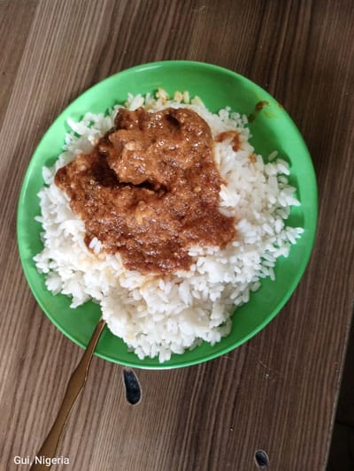 An overhead shot presents a meal served on a round, bright green plastic plate, positioned on a brown wooden table with visible grain patterns. The plate holds a generous portion of white, cooked rice, mounded and somewhat unevenly distributed. A thick, reddish-brown stew or sauce heavily covers the top and central area of the rice, featuring chunks of what appears to be meat or protein within its textured surface. Small splatters of the reddish-brown sauce are visible on the green rim of the plate and scattered lightly on some of the exposed rice grains. A golden-colored metallic spoon is partially visible at the bottom of the image, resting on the wooden table, with its handle extending towards the plate. Text overlay in the bottom left corner of the image reads "Gui, Nigeria". The scene is located in the city of Kuje, Nigeria.