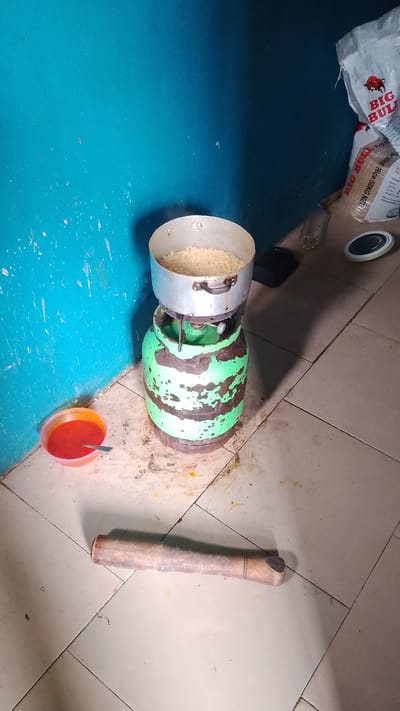 In the city of Asubi, Nigeria, a domestic scene unfolds in a room with turquoise-painted walls and a tiled floor. A green and black striped gas cylinder serves as a makeshift stove, supporting a metal pot filled with white rice. To the left, a small plastic bowl contains a vibrant red sauce and a spoon. In the foreground, a wooden rolling pin lies on the floor. In the background, a white bag with red lettering, likely containing rice, is leaning against the wall, and a plastic bottle and a dark object are also visible. The scene suggests food preparation is in progress.