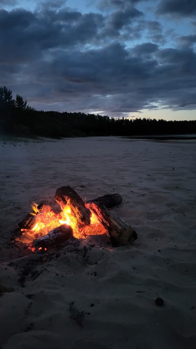 The image shows a campfire burning on a sandy beach at dusk. The sky is filled with dark, dramatic clouds, hinting at an approaching storm or the end of daylight. The fire is central to the composition, with logs burning and emitting a warm, orange glow. The sand around the fire is illuminated by its light, showing subtle ripples and textures. In the background, a dark line of trees forms a silhouette against the twilight sky, and beyond that, a body of water is visible. The overall atmosphere is one of a serene yet moody evening by the water. The location is specified as Zaostrov'e, Russia. There are no people visible in the image.