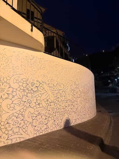 At night in Ponta do Sol, Portugal, a bridge or walkway is adorned with a mural of flowers. The mural is drawn in blue lines on a white background, with the floral pattern fading towards the left side of the structure. The structure itself is adjacent to a building with an exterior staircase. To the right of the walkway is a raised stone path or wall. The sky is dark, with some lighting visible in the distance.