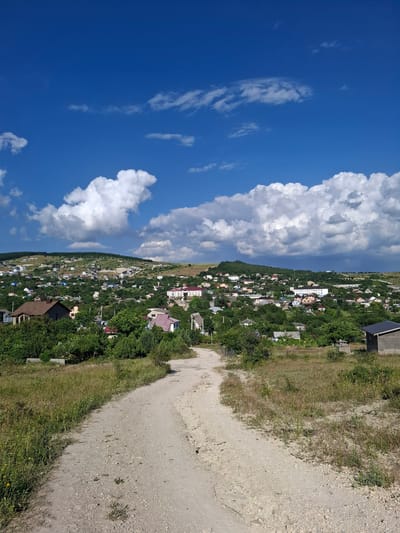 This image depicts a view of the city of Varvarovka, Russia, on a sunny day with a partially cloudy sky. In the foreground, a dirt road curves into the scene from the bottom right, bordered by dry grass and sparse vegetation. The road leads towards a hillside densely populated with houses and trees. The houses vary in size and architectural style, with many featuring red or gray roofs. Some larger buildings, possibly community structures or apartment complexes, are visible on the upper part of the hill. The sky is a vibrant blue, with large, fluffy white clouds scattered across it. The overall impression is one of a rural or semi-rural settlement nestled within a hilly landscape. There are no people visible in the image.