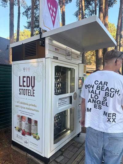 This media file captures a young person, viewed from behind and from the side, standing in front of a modern, white ice cream vending machine. The person is wearing a white t-shirt with large black text that reads "CAR POV BON BEACH, VALI BU LOS ANGEL ES M MER XX". They are also wearing light blue jeans.
The vending machine is the central object, displaying an array of ice cream products within its glass front. The machine has prominent branding with "EIS Berger" logos and text in Latvian that translates to "ICE CREAM STORE". A large graphic on the lower part of the machine advertises "0% artificial additives. 100% pure pleasure." and lists various flavors like "Tahiti Vanilla", "Strawberry Balsamic", and "Sicilian Pistachio". There is also a QR code and a website address: www.ledustotele.lt.
The setting appears to be outdoors, with tall pine trees visible in the background, suggesting a natural environment like a park or a forest. The ground is paved with brick-like stones in some areas. Sunlight is present, indicating it's daytime, possibly late afternoon or early evening given the angle of the light. The overall scene conveys a casual, everyday moment of someone about to purchase or having just purchased ice cream from a high-tech vending machine.