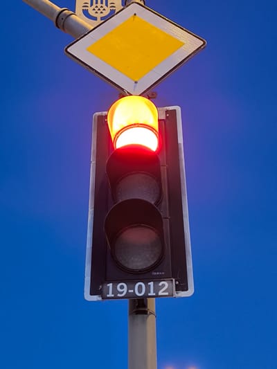 This media file shows a traffic light against a clear, deep blue sky, suggesting it is either early morning or late evening. The red light is illuminated, indicating a stop. Above the traffic light, a diamond-shaped yellow sign with a white border is mounted, which is a common road sign indicating a priority road or right of way. Below the traffic light, a rectangular plate with white text "19-012" is visible, likely an identifier for the traffic light. The traffic light itself is mounted on a metal pole, which is also visible in the frame. The overall scene is set in Kartal, Turkey, based on the provided location context. There are no people visible in the image. The weather appears clear.