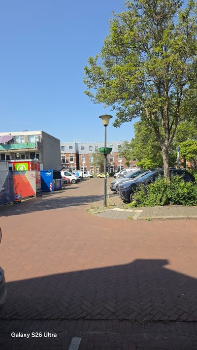 This image depicts a street scene in Vlaardingen, Netherlands. The perspective is from a slightly elevated angle, looking down a brick-paved sidewalk. On the left side of the image, a large tree with lush green leaves dominates the upper portion, casting a shadow onto the sidewalk. A lamppost with a cream-colored shade stands near the tree.
Behind the tree and lamppost, apartment buildings are visible. These buildings appear to be multi-story structures with modern architecture, featuring white and grey facades with numerous windows. Several cars are parked along the side of the street, including a dark-colored SUV, a white car, and a few others further down.
In the foreground, towards the right, the brick pavement is clearly visible, with some darker asphalt sections and grass growing between the bricks. The shadow of an unseen object, possibly a person or structure, falls across the sidewalk, indicating it is daytime with a clear sky, likely late afternoon given the long shadows.
Towards the bottom right of the image, the text "Galaxy S26 Ultra" is watermarked, suggesting this is a photograph taken with a Samsung Galaxy S26 Ultra phone. There are also some large blue and red waste bins visible near the buildings, indicating a residential or mixed-use area. No people are clearly visible in the image. The overall impression is a quiet, ordinary day in a European urban environment.