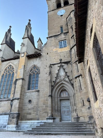 The image captures a low-angle exterior view of a stone church with Gothic architectural elements in Saint-Antoine-l'Abbaye, France. The building features a tall clock tower with a clock face displaying roman numerals, indicating the time is approximately 10:50. Two prominent pointed spires with crosses atop them rise from the left side of the church, reaching towards a clear, pale blue sky. The facade of the church is constructed from weathered, light-colored stone blocks. Intricate tracery adorns large Gothic windows with diamond-shaped panes. A grand, arched doorway with a weathered, light blue wooden door is visible at the base of the central section of the church, accessible by a wide set of stone stairs. To the right of the main church structure, another stone building with a textured facade and barred windows is partially visible. The ground in front of the church is covered with fine gravel. The overall impression is one of historical grandeur and architectural detail.