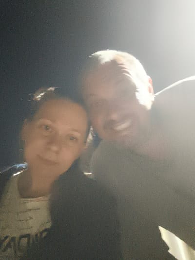 The image is a blurry, low-resolution selfie taken at night, possibly outdoors. Two individuals are visible. On the left is a woman with dark hair pulled back, looking towards the camera with a slight smile. She is wearing a light-colored top with some text on it. On the right is a man, facing slightly away from the camera but with his head turned to reveal his face. He has fair hair and is smiling broadly. The background is dark, with a bright light source, possibly a streetlamp or flashlight, creating a strong glare that washes out details in the upper right portion of the image. No specific landmarks of Il'mino, Russia, are discernible.