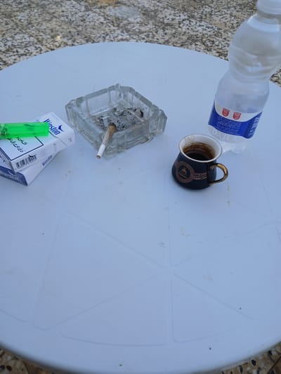 The image shows a white plastic table outdoors, with a cobblestone-like ground visible at the bottom. On the table are several items: a clear glass ashtray with cigarette butts and ash, a green lighter, a pack of Winston cigarettes, a bottle of water, and a small dark cup filled with coffee. The coffee cup has a gold rim and a decorative gold pattern. The water bottle label is partially visible, showing "DAJLA" and some text in Arabic. The lighting suggests it is daytime. The overall scene depicts a casual outdoor setting, possibly a cafe or patio.