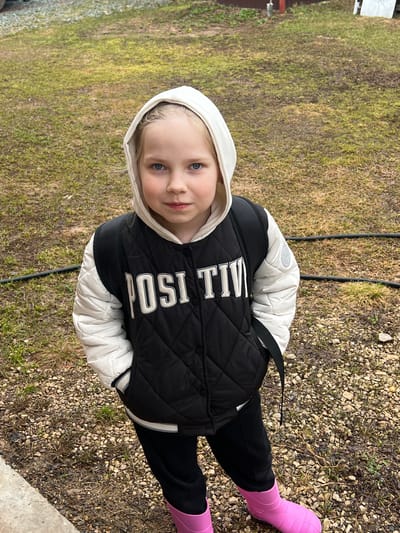A young child with fair skin, blue eyes, and blonde hair stands outdoors. The child wears a white hooded jacket with a black quilted vest over it, the vest featuring the word "POSITIVE" in white block letters. The child also wears black pants and bright pink rubber boots. The ground consists of grass and gravel. The location is Bol'shakino, Russia.