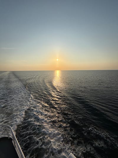 The image depicts an open body of water under a clear sky, with the sun low on the horizon, creating a sunrise or sunset scenario. The location is consistent with an oceanic or large sea environment near Kato Karioutes, Greece, as specified.
**Subjects and Objects:**
- **Sky:** The upper two-thirds of the frame are occupied by the sky, which transitions from a light, pale blue at the zenith to a warm, soft yellow-orange at the horizon line.
- **Sun:** A distinct, bright yellow-orange spherical object, interpreted as the sun, is positioned just above the horizon, slightly to the right of the frame's horizontal center.
- **Water:** The lower third of the frame comprises the surface of the sea, exhibiting a dark blue-grey hue. The water shows subtle rippling patterns across its expanse.
- **Sunlight Reflection:** A prominent vertical band of shimmering golden light extends from the sun's position across the water towards the foreground, creating a bright path on the water's surface.
- **Vessel Wake:** In the left half of the image, a wide, turbulent trail of white foam, consistent with a vessel's wake, extends diagonally from the lower left corner towards the mid-left of the frame. This wake contrasts sharply with the darker water and intersects with the reflective path of sunlight.
- **Vessel Structure:** A segment of a white railing or deck structure from a vessel is visible in the extreme lower-left corner of the frame, indicating the photograph was taken from aboard a moving watercraft.
**Actions and Interactions:**
- The sun is either rising or setting, casting a warm glow across the sky and reflecting brightly on the water's surface.
- A vessel is actively moving forward, indicated by the trailing wake, which generates disturbed, frothy water.
- Light from the sun interacts with the water, creating a specular reflection path.
**Environment:**
- The environment is an expansive open sea with no visible landmasses, under calm atmospheric conditions. The lighting indicates either early morning or late evening.