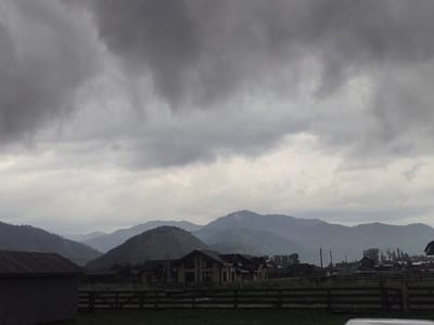 The image depicts a panoramic view of a rural landscape under a heavily overcast sky. In the foreground, to the left, is the side of a grey building with a dark, sloped roof. A wooden fence runs horizontally across the mid-ground, separating the foreground from a cluster of modern houses with dark roofs, situated in front of rolling hills. The houses appear to be of similar architectural style, with visible windows and dark trim. Beyond the houses, a series of layered, green mountains recede into the distance, shrouded in atmospheric haze. The overall impression is one of a calm, possibly cool, day in a mountainous region. The scene is identified as being in the city of Anos, Russia. There are no people or animals visible in the image.