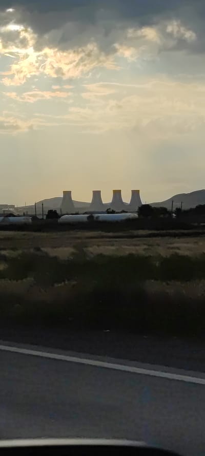 The image shows a landscape scene with several cooling towers from a power plant in the distance, silhouetted against a hazy, overcast sky. The cooling towers are tall and conical, with yellow tops. In the foreground, there is a field with some vegetation and what appear to be greenhouses. A road with white lines is visible in the bottom part of the image. The overall impression is of an industrial area situated in a rural or semi-rural setting. The location is identified as Haytagh, Armenia.