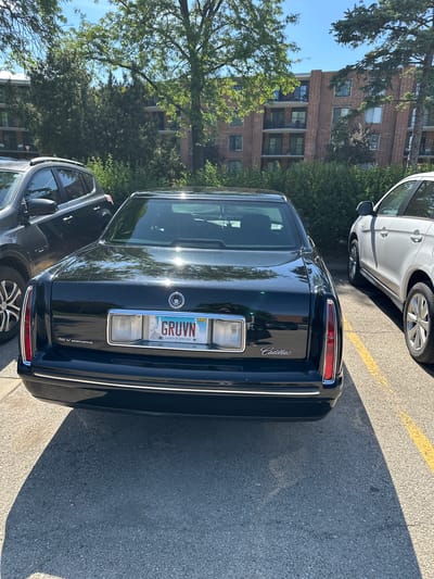 A black Cadillac sedan is parked in a parking lot with yellow lines, alongside a white SUV and a gray SUV. The Cadillac's license plate reads "GRUVN" and indicates it is from the "Land of Lincoln," Illinois. The Cadillac also features "Cadillac" script on the right side of the trunk and "32 V Northstar" on the lower left side. In the background, trees and a brick building are visible. The photograph was taken in Arlington Heights, Illinois, USA.