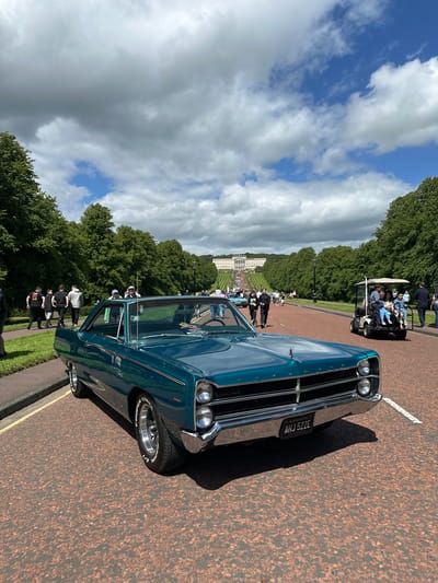 This is a full-shot, eye-level, outdoor photograph of a vintage teal-colored Plymouth Fury parked on a red asphalt road. The car is facing slightly to the right of the camera. The license plate reads "AHJ 522E." In the background, a large white building with columns and a dome is visible, situated at the top of a wide, tree-lined avenue. This is the Stormont Estate, home to the Parliament of Northern Ireland, located in Belfast, United Kingdom. Several people are walking on the left side of the avenue, and a golf cart carrying passengers is driving on the right side of the road. The sky is partly cloudy with a mix of white and dark gray clouds. The overall scene suggests an outdoor event or gathering, likely a car show given the presence of the vintage vehicle.
