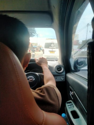 The image shows the interior of a car, with the focus on the driver's right hand on the steering wheel and the back of the driver's head. The driver is wearing a brown t-shirt. The car is in motion, as indicated by the blurred background and the open window with a side-view mirror. Outside the car, a street scene is visible, with buildings, power lines, and other vehicles. Specifically, a white van with a license plate that appears to be "BL 7572 AB" is in front of the car. A motorcycle with a rider is also seen on the road. The environment suggests a semi-urban or rural setting in Indonesia, potentially in the city of Damatutong, based on the provided location information. The lighting suggests daytime.