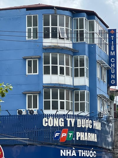 The image shows a blue, multi-story building with a series of windows. A blue sign with white text is attached to the right side of the building. The sign reads "TIEM CHUNG" in vertical orientation. Below the building, a blue fence is visible with white lettering that says "CONG TY DUOC PHAM" and "FPT PHARMA". Another blue sign with white lettering below this reads "NHA THUOC". There are also two air conditioning units mounted on the exterior of the building above the fence. Power lines are strung across the scene. The sky is overcast. The location is specified as Hà Giang 2 Ward, Vietnam.