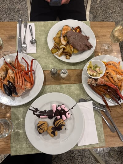 The image captures a top-down view of a dinner spread on a wooden table, possibly at a restaurant in Es Camp De Mar, Spain, given the context. The table is partially covered with a muted green placemat.

The main subjects are plates of food. One plate features a generous portion of cooked seafood, including large prawns, crab legs, and mussels, arranged over what appears to be more prawns. Another white plate holds a slice of roasted meat, served with roasted potatoes and onions. A third plate contains a dessert assortment: pink marshmallows drizzled with chocolate sauce, and small cream puffs also topped with chocolate. A small bowl of salad with cheese and vegetables is positioned near the seafood.

Accompanying the food are various objects. Two salt and pepper shakers are visible. Cutlery, including forks, knives, and spoons, is laid out on napkins. A glass of white wine, partially filled, sits on the table, along with another glass which is mostly out of frame. The presence of multiple plates and cutlery suggests a meal is either in progress or about to begin, likely a celebratory or a leisurely dinner.

There are no visible people in the frame. The lighting suggests it could be daytime, possibly late afternoon or evening, given the indoor setting and the available light. The overall atmosphere appears relaxed and convivial, centered around enjoying a diverse meal. No text is discernible in the image.