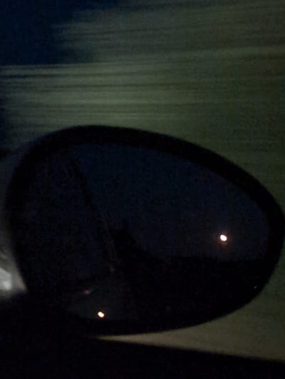 The image shows a car's side mirror reflecting a dark, hazy scene. In the reflection, a bright, crescent-shaped light is visible, possibly the moon or a distant streetlight. The surrounding environment in the reflection appears to be a dark, possibly rural or semi-rural area with horizontal lines suggesting clouds or a distant horizon. The car is likely moving, as indicated by the motion blur in the background. The location is Courtisols, France, but no specific landmarks are identifiable. The image is grainy and dark, making detailed analysis challenging.