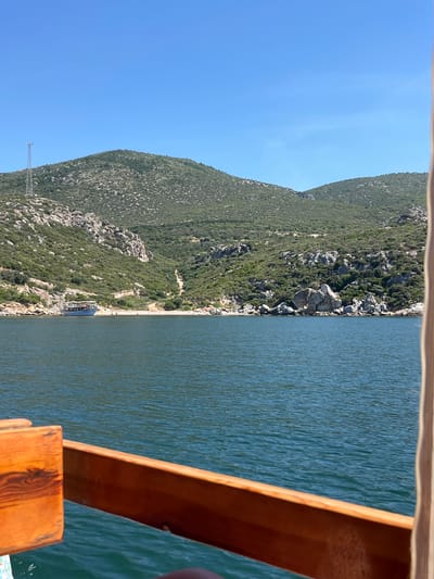 The image, correctly oriented, presents a vibrant daytime scene captured from the perspective of a boat on a calm body of water.

In the immediate foreground, a warm-toned wooden railing or structure of the boat from which the photo was taken frames the lower right portion of the image. Beyond this, a wide expanse of deep blue-green water stretches across the frame, showing subtle ripples on its surface, indicating calm conditions.

The left side of the image is dominated by a majestic coastline, characterized by steep, rocky hills covered in dense green vegetation, including shrubs and trees. The hills rise dramatically from the water's edge. Along the base of these hills, a narrow, light-colored strip, possibly a path or a small beach, is visible. Further along this shore, a white and blue boat, likely a passenger or tour vessel, is docked or anchored close to the land. High on the hillside, against the clear blue sky, a metallic lattice structure, possibly a communication tower, stands out.

The sky is a brilliant, cloudless blue, suggesting clear weather and ample sunshine. No distinct people are visible, and there is no discernible text in the image. The overall setting depicts a serene coastal environment, typical of areas like Menderes, Turkey, suggesting a leisurely boat trip or a peaceful observation of the natural landscape.