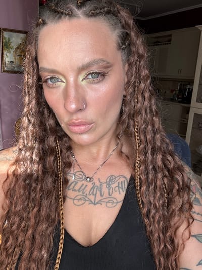 This is a portrait-style, medium shot of a young woman lying on her side. She has long, curly, dark brown hair with lighter brown braided accents. Her face is turned towards the camera, and she has striking blue eyes with pale green eyeshadow. She is wearing a silver necklace with a circular pendant and a small zipper charm. She has a nose ring and a lip piercing. There is a tattoo visible on her chest and upper arm, with stylized lettering. The background appears to be an indoor setting with cabinets or shelves visible in the upper portion of the frame, and a framed picture on the wall in the lower left. The lighting suggests it might be daytime, with some artificial light source contributing to the overall illumination. The mood is somewhat relaxed or contemplative. There is no visible text in the image.