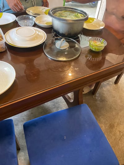 The image depicts a dining table laden with dishes, a pot, and serving bowls. A person is partially visible on the left side of the frame, along with their hand. A person with visible skin and clothing is at the top left of the frame. The table has a glossy brown surface and is surrounded by at least two blue upholstered chairs. The location is Hijo, Philippines.