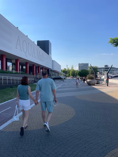A couple is walking hand-in-hand on a paved walkway in front of a modern building complex that reads "AQUA Mall" in large letters. The woman, on the left, is wearing a green sleeveless top and white shorts. The man, on the right, is wearing a light blue t-shirt and matching shorts. He is holding a bottle of water in his right hand. The walkway is made of patterned pavers, with a red and white striped bike path running alongside it to the left. In the background, beyond the Aqua Mall, there are more buildings, trees, and a body of water with boats docked. The sky is clear and blue. The scene appears to be in a waterfront area of a city. The location is Maiorovskii, Russia.
