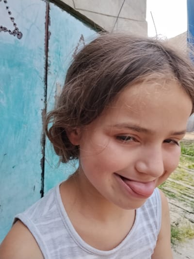 The image shows a close-up of a young girl with light brown hair, wearing a light gray tank top. She is outdoors, with a bright blue, weathered metal door to her left and a blurred background of greenery and what appears to be wooden planks. The girl is looking down and to her right, with a playful expression, sticking her tongue out slightly. Her eyes are dark, and her skin is fair with rosy cheeks. The lighting suggests it is daytime, likely outdoors. The setting appears to be informal, possibly a backyard or a rural area, consistent with the location context of Vulcănești, Moldova. There is no visible text in the image.