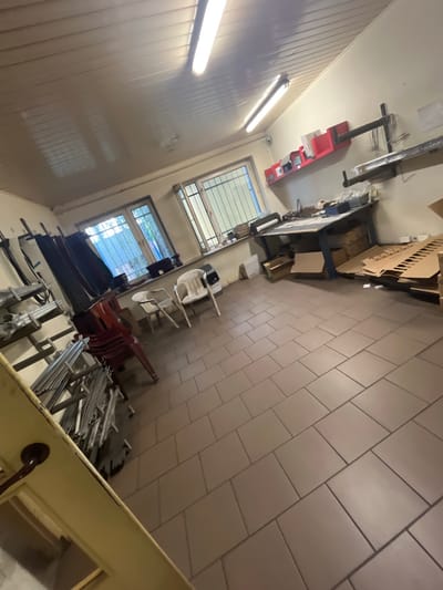 This is a medium-shot, interior photograph of a workshop or storage area. The room is rectangular with light brown tiled flooring. The walls are a neutral off-white color. The ceiling is paneled with two long fluorescent light fixtures illuminating the space.

In the foreground, to the left, a light yellow door is partially visible, with a dark brown handle. A stack of metal materials is organized on shelves to the right of the door.

The middle ground features a large workbench with a blue metal frame and a wooden top, cluttered with various items including boxes, paper rolls, and what appears to be a stapler or labeling machine. To the right of the workbench, there are stacks of flattened cardboard boxes. Further to the right, a tall shelf unit holds more boxes and materials.

In the background, there are two windows with metal grating, suggesting security. The left window has blue curtains. Three red plastic chairs are stacked to the left of the windows, and two white plastic chairs are placed in front of the right window. A red shelf unit with compartments is mounted on the wall above the workbench, holding documents or files.

The overall impression is of a functional, somewhat disorganized workspace. The location is identified as a workshop in Bogucin, Poland.
