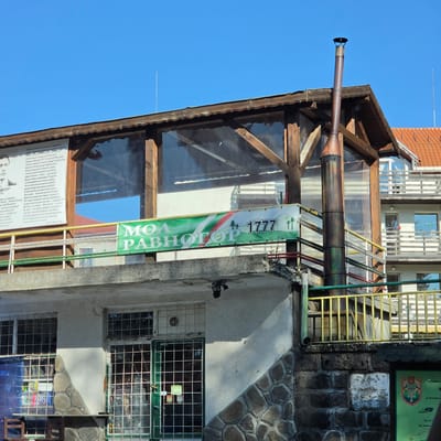The image depicts an outdoor scene in Ravnogor, Bulgaria, featuring a two-story building with a rustic wooden and glass exterior on the upper level and a stone-clad lower level. A prominent, vertical chimney pipe, exhibiting a distressed metallic finish, rises from the side of the upper structure. A banner, predominantly green with white and red accents, is stretched across the railing of the upper level, displaying the text "MОЯ РАВНОГОР" (My Ravnogor) and the number "1777", along with stylized tree graphics. Above the banner, a large, framed text is affixed to the building, with a black and white portrait of a man on the left side. The lower level has barred windows and a green-framed door. The surrounding environment suggests a sunny day with a clear blue sky. Additional buildings, including one with an orange tiled roof, are visible in the background. A security camera is mounted beneath the overhang of the lower level.