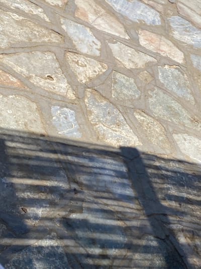 The image depicts a sunlit outdoor paved surface, likely a walkway or patio, composed of irregularly shaped flagstones or natural stone tiles in various shades of grey, beige, and light brown, set with visible mortar joints. The upper two-thirds of the frame show the sun-drenched pavement. The lower one-third of the image is covered by distinct shadows. A prominent, elongated shadow resembling a human form, with a discernible head-like shape, is cast from the right side towards the lower center. Overlapping this and extending across the lower half are multiple parallel, horizontal bar-like shadows, consistent with those cast by a railing or fence structure. There are no people, objects, or actions other than the interplay of light and shadow on the pavement. The precise location within the city of Φάρος, Greece, cannot be determined from the visual information provided.