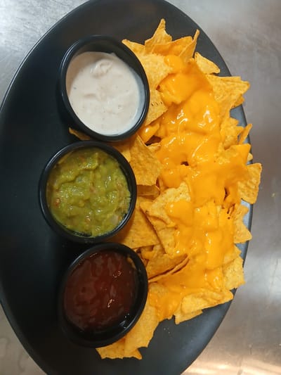 The image captures a visually appealing platter of nachos, generously prepared on a black, oval-shaped serving dish. A large pile of golden-brown tortilla chips forms the base, completely covered in a thick, vibrant orange melted cheese sauce, which drips enticingly over the chips.

Three small, black, round ramekins containing various dips are arranged at the bottom edge of the plate. From left to right: the first ramekin holds a creamy, off-white sauce, likely sour cream, ranch, or a similar dairy-based dip. The middle ramekin contains a chunky, bright green guacamole, showing discernible pieces of avocado and possibly other ingredients. The third ramekin on the right holds a reddish-brown sauce, which appears to be a salsa or a sweet chili sauce, characterized by its glossy texture.

The setting suggests a food service environment, possibly a kitchen or restaurant in Sotira, Cyprus, given the metallic, slightly scratched surface visible in the background. This surface reflects ambient light, indicating a well-lit area. The dish is presented as ready for immediate consumption, emphasizing its freshness and appeal. There are no people, visible text, or discernible time of day or weather elements in the image.