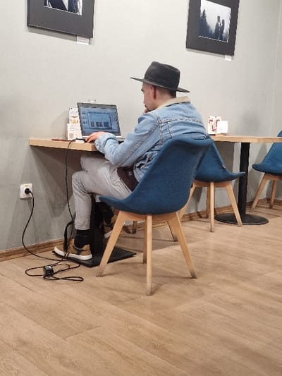 An interior scene captured in Odorheiu Secuiesc, Romania, shows a male individual seated at a light-colored wooden table, engaged with a laptop. The subject, viewed from the rear, wears a dark fedora-style hat, a light-blue denim jacket with a cream-colored fleece collar, light-grey trousers, and tan sneakers with dark accents. His left hand rests on the laptop's keyboard, while his right hand is positioned near his chin.
The silver-colored laptop displays a graphical interface with multiple windows open. To the left of the laptop, a power cord extends from a white adapter plugged into a black wall outlet on the light-grey wall, connecting to the laptop. Adjacent to the laptop, a small white stand holds brochures or menus, one displaying a visible QR code. Further right on the table, a small, pink-wrapped object is present.
The subject occupies a blue fabric chair with light-colored wooden legs. A black, low-profile footrest is positioned beneath his feet. To the right, another similar blue chair and a table with a dark, circular base are visible. Two framed black-and-white photographs adorn the wall, each with a small white label beneath. The flooring consists of light-colored, wood-patterned planks.