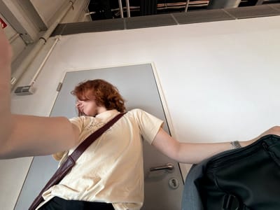 A person with red hair, wearing a tan t-shirt and carrying a brown shoulder bag, is standing in what appears to be an indoor space, possibly a building or public transportation vehicle. The scene is framed with elements such as a gray door with a silver handle, a black backpack and a gray garment, and architectural details including ceiling vents and pipes. The location is Lautzenhausen, Germany.